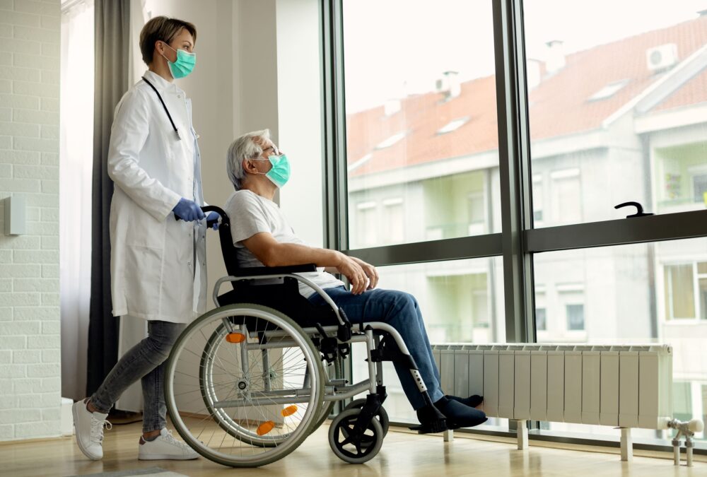 Um acompanhante de hospital profissional da Pronto Care, uniformizado em azul, segura e conforta a mão de um paciente idoso deitado na cama de hospital, oferecendo suporte emocional durante a internação.