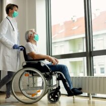 Um acompanhante de hospital profissional da Pronto Care, uniformizado em azul, segura e conforta a mão de um paciente idoso deitado na cama de hospital, oferecendo suporte emocional durante a internação.