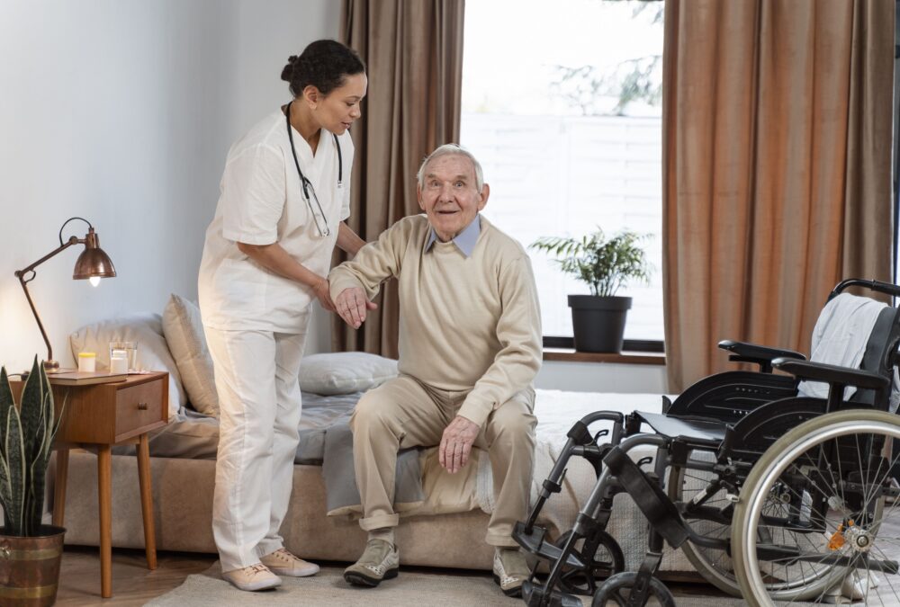 Um profissional de assistência domiciliar da Pronto Care, vestindo uniforme branco e estetoscópio, auxilia com cuidado um idoso sorridente a se levantar de uma cama confortável ao lado de uma cadeira de rodas.