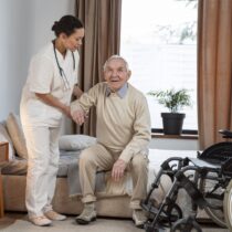 Um profissional de assistência domiciliar da Pronto Care, vestindo uniforme branco e estetoscópio, auxilia com cuidado um idoso sorridente a se levantar de uma cama confortável ao lado de uma cadeira de rodas.